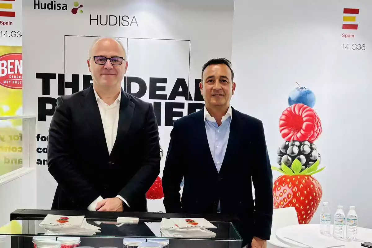 Dos hombres de pie en un stand de exhibición de Hudisa, con un fondo que muestra frutas y el logotipo de la empresa.