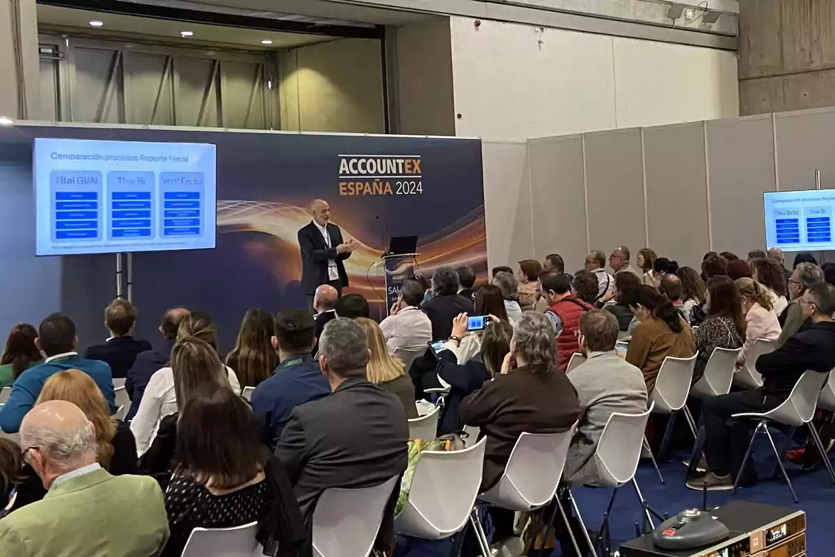 Una conferencia en Accountex España 2024 con un presentador hablando frente a una audiencia atenta y una pantalla mostrando información sobre procesos de reporte fiscal.