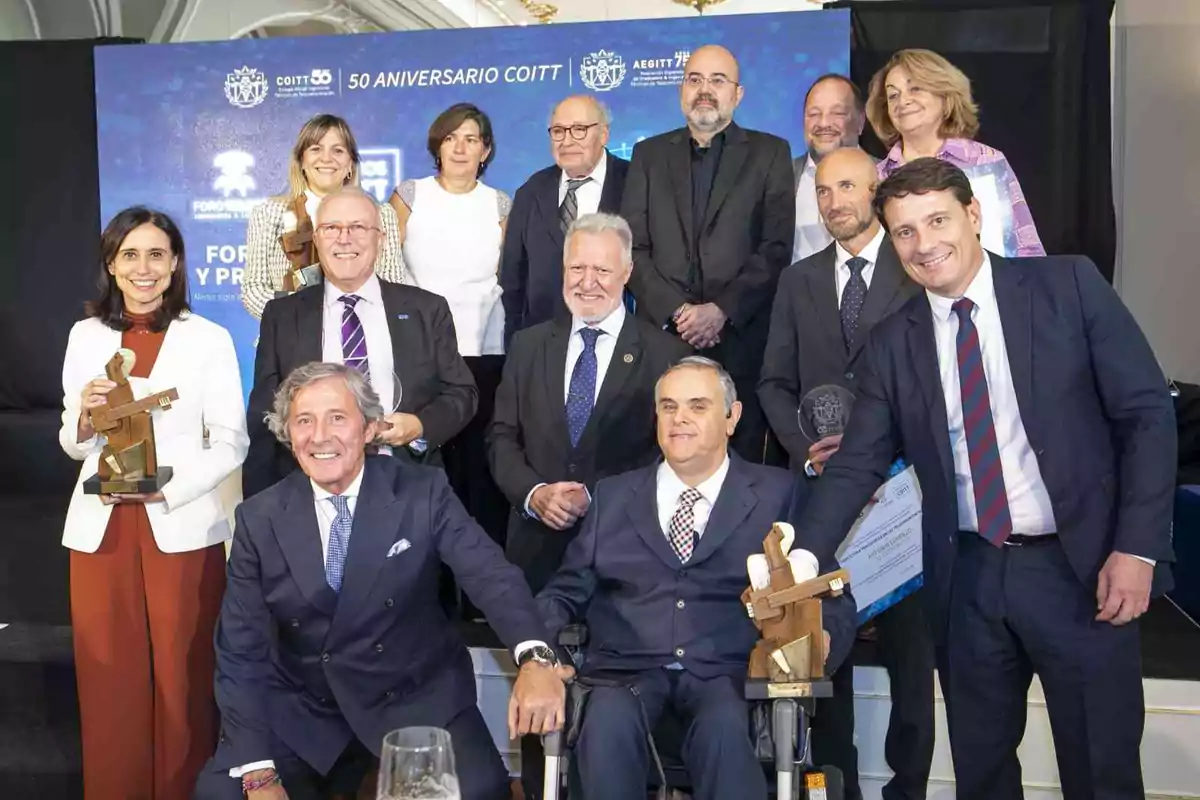 Un grupo de personas vestidas formalmente posan para una foto en un evento de aniversario, algunas sostienen trofeos y certificados.