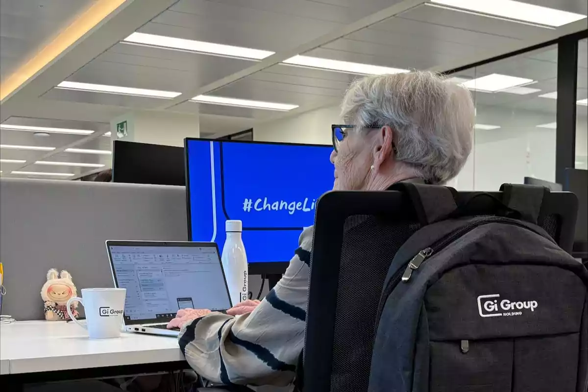 Mujer mayor de cabello canoso sentada en una oficina moderna trabajando en una laptop con una mochila negra de Gi Group en la silla y una pantalla azul al fondo que muestra el texto #ChangeLi
