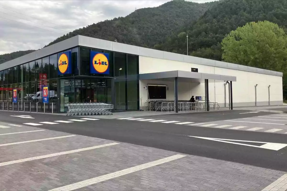 Un supermercado Lidl con un diseño moderno y un estacionamiento vacío, rodeado de montañas y vegetación.