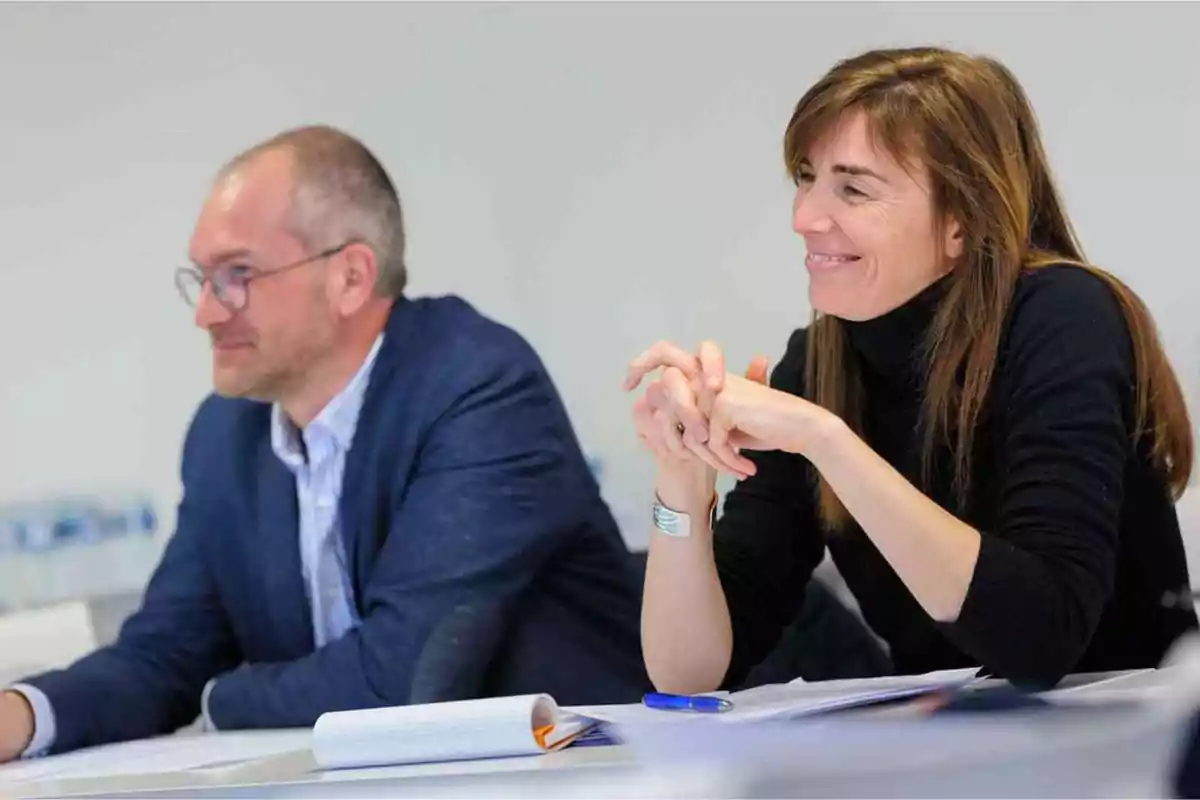 Dos personas sentadas en una mesa durante una reunión, una mujer sonríe mientras escucha atentamente.