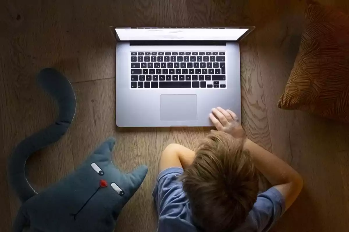 Niño acostado en el suelo usando una laptop junto a un cojín en forma de gato y un cojín naranja