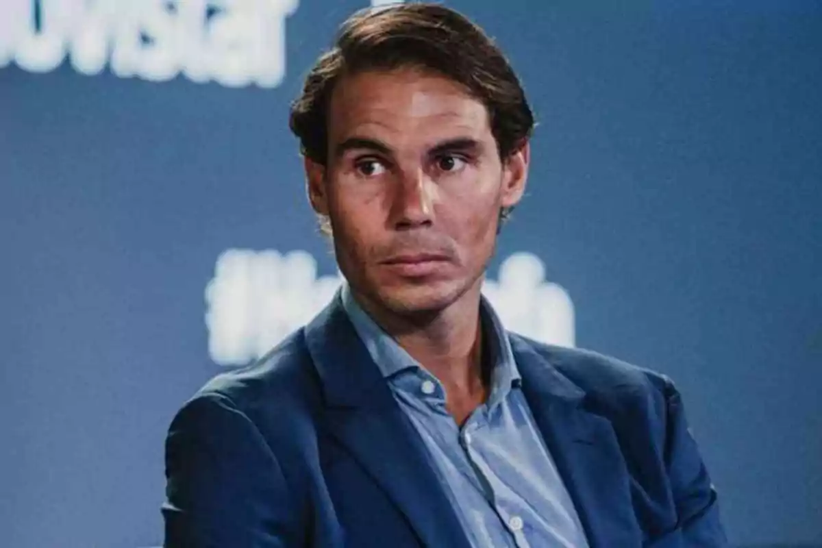 Un hombre con traje azul y camisa clara mira hacia un lado con expresión seria frente a un fondo azul.