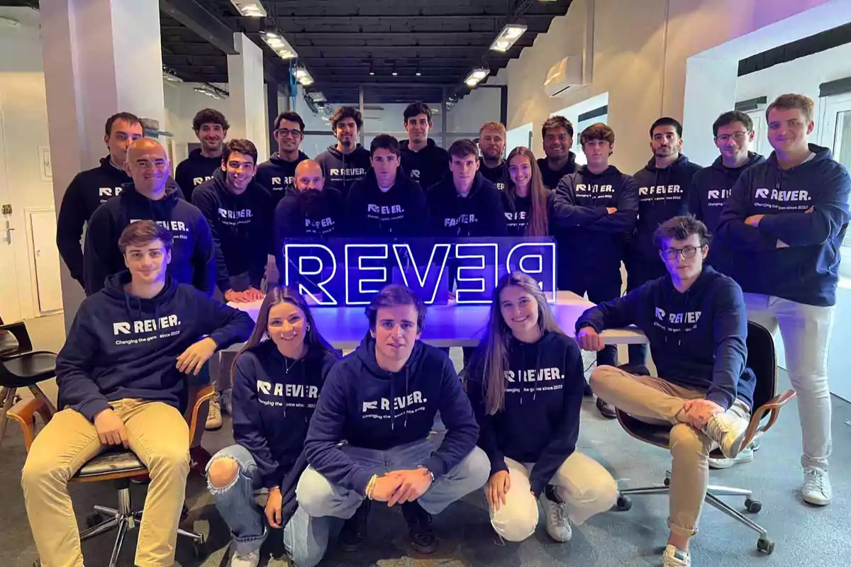 Un grupo de personas posando juntas en una oficina, todas llevan sudaderas con el logo de "REVER" y están alrededor de un letrero iluminado con el mismo nombre.