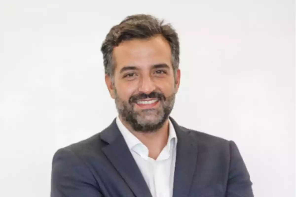 Hombre de cabello y barba oscura con saco azul y camisa blanca sonriendo frente a un fondo claro