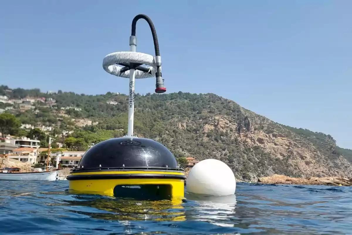 Un dispositivo flotante de monitoreo en el agua con un paisaje montañoso y casas en el fondo.