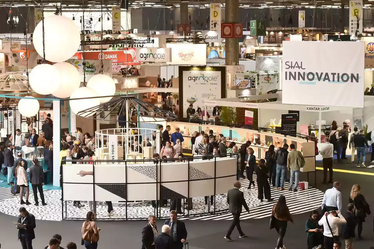 Una feria comercial con numerosos stands y personas interactuando en un espacio amplio y bien iluminado.