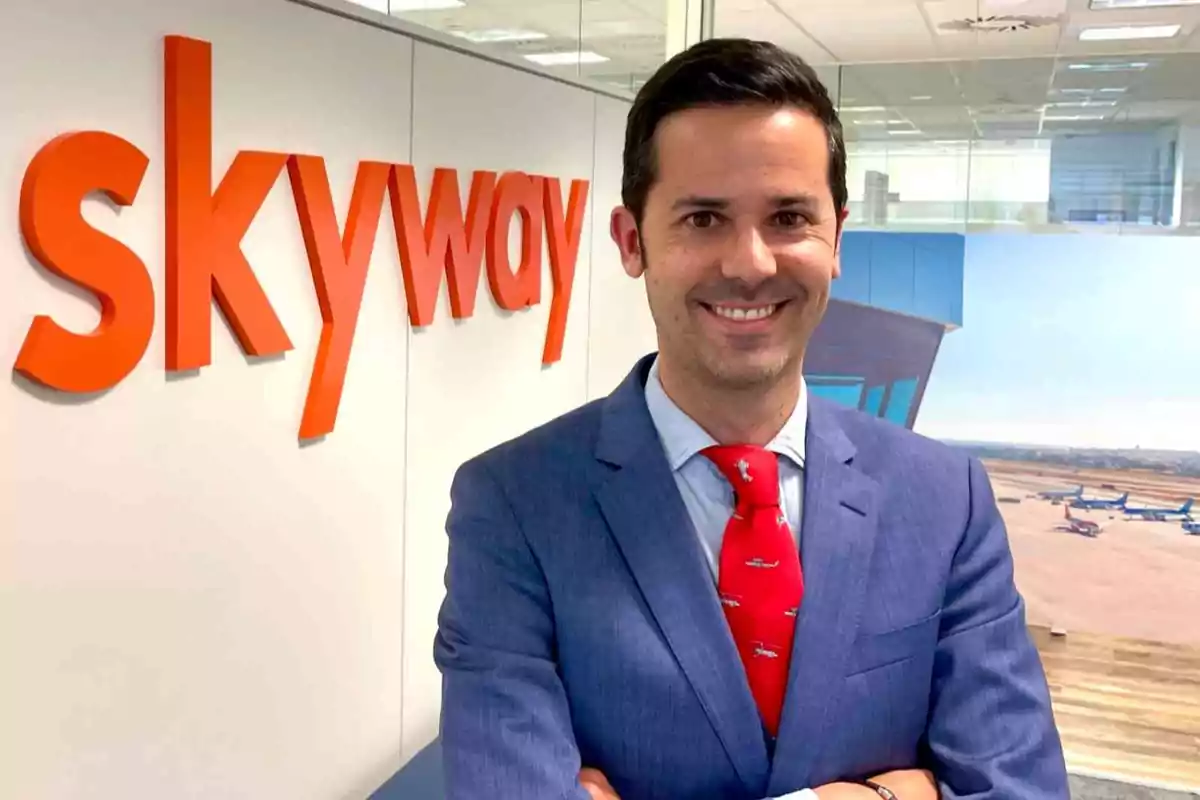 Hombre de traje azul y corbata roja sonríe frente a una pared con el logo de skyway en una oficina moderna