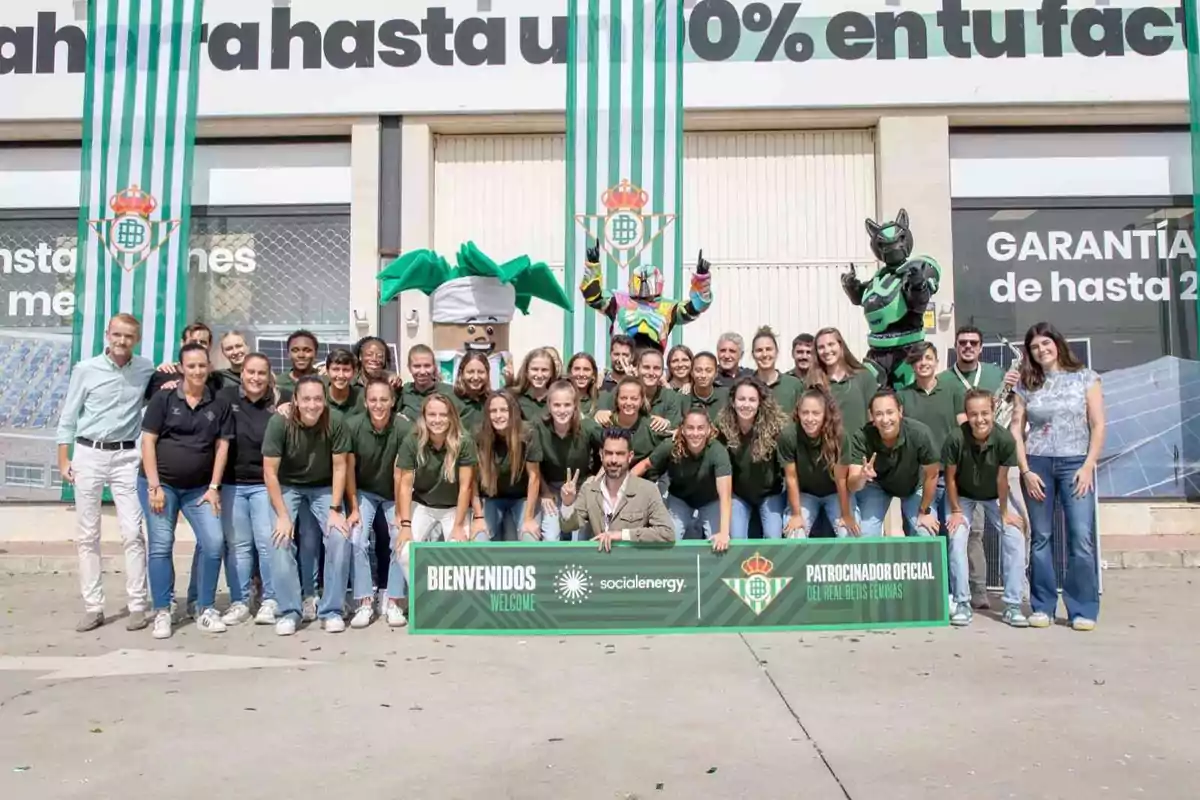 Un grupo de personas posando frente a un edificio con una pancarta que dice "Bienvenidos" y "Patrocinador Oficial del Real Betis Féminas", con dos mascotas y banderas verdes y blancas.