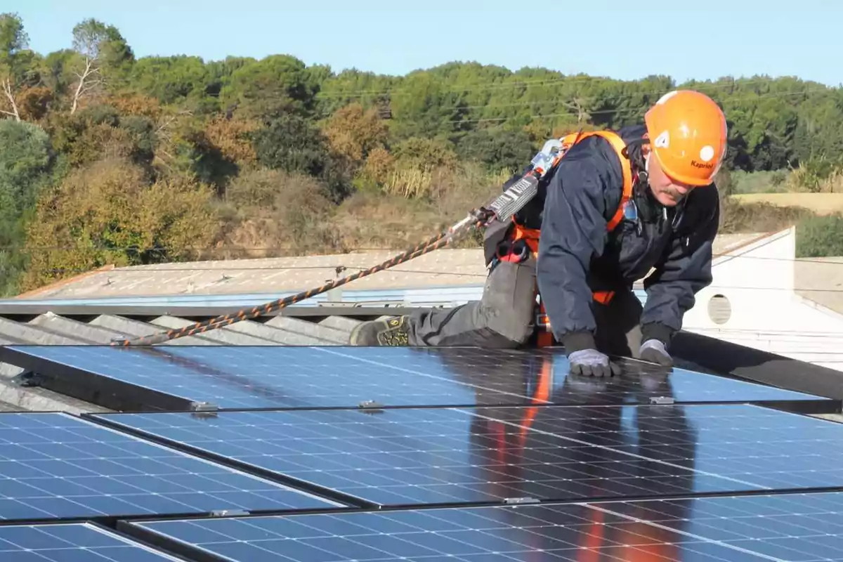 Trabajador con casco y arnés instalando paneles solares en un techo con árboles al fondo.