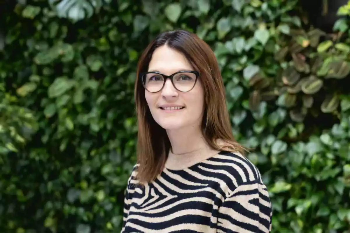 Mujer de cabello castaño y gafas sonríe frente a un fondo de plantas verdes, lleva un suéter de rayas tipo animal print.