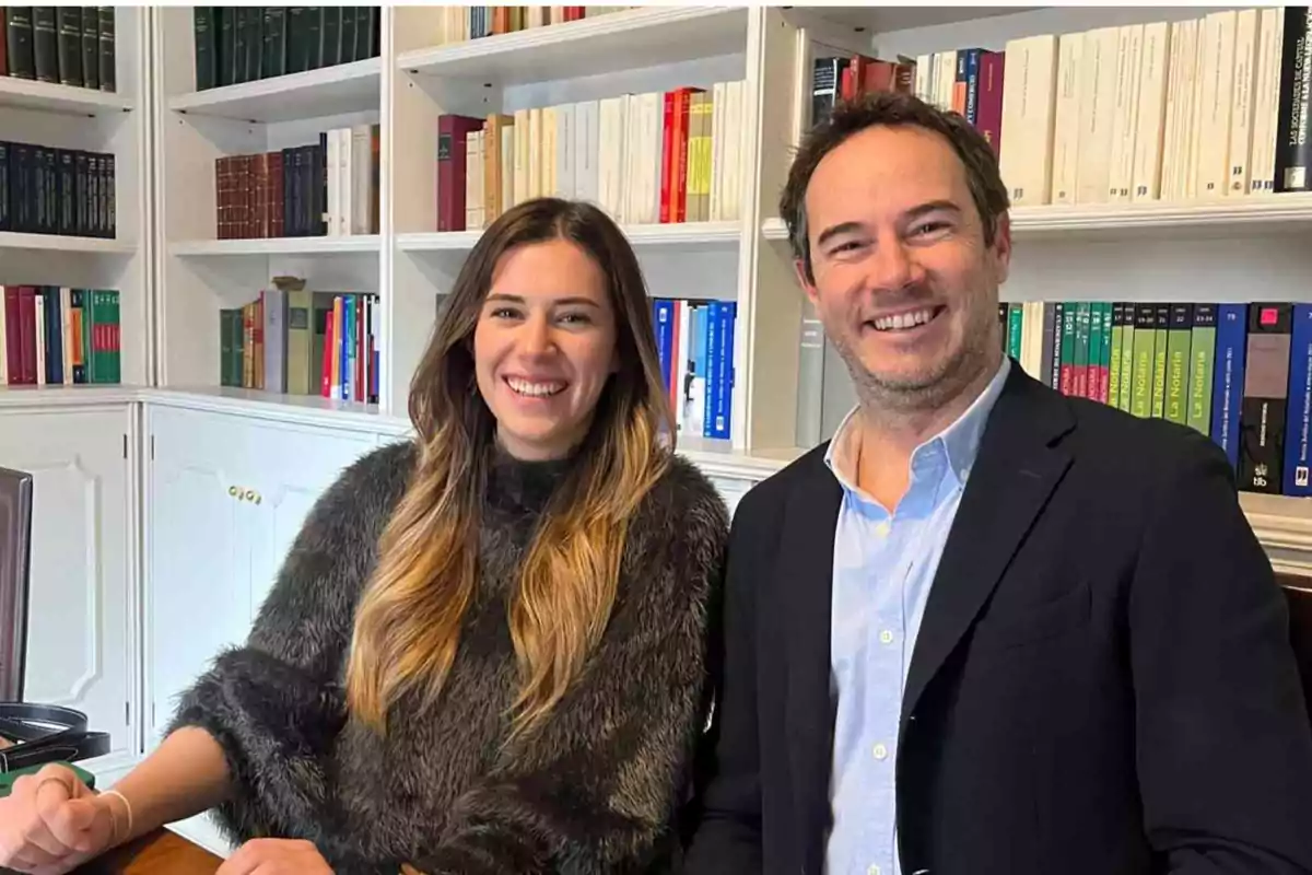 Dos personas sonrientes sentadas en una oficina con estanterías llenas de libros al fondo.