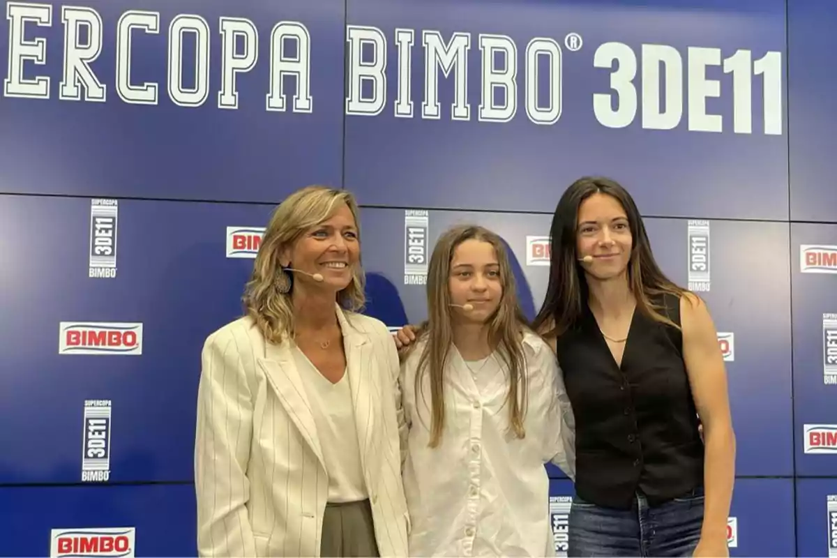 En la imagen se puede ver a tres personas posando frente a un fondo azul que tiene el logotipo de "BIMBO" y el texto "SUPERCOPA BIMBO 3DE11". Las tres personas están sonriendo y llevan micrófonos de diadema, lo que sugiere que están participando en un evento o presentación. La persona a la izquierda lleva un traje claro, la persona en el centro lleva una camisa blanca y la persona a la derecha lleva una camiseta sin mangas negra.