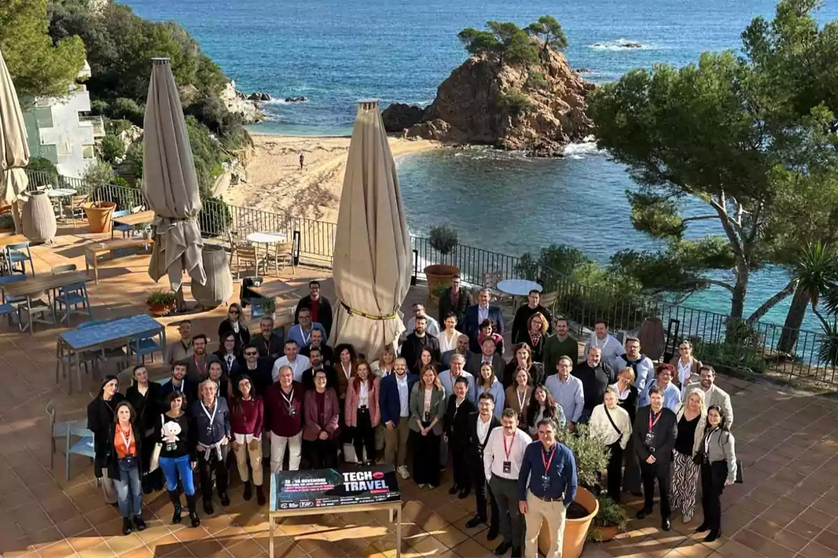 Un grupo de personas posando en una terraza con vista al mar y una pequeña isla rocosa al fondo, rodeados de mesas y sombrillas cerradas.