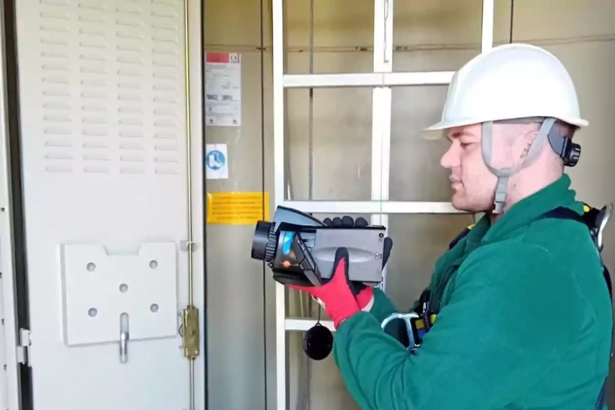 Trabajador con casco y guantes rojos utilizando un dispositivo de medición frente a un panel eléctrico.