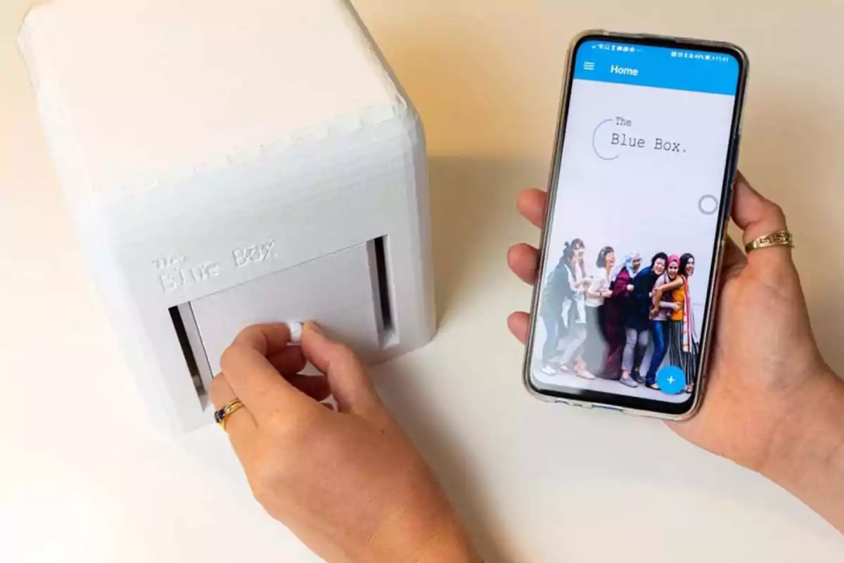 Persona sosteniendo un teléfono móvil con la aplicación "The Blue Box" y manipulando un dispositivo blanco rectangular.