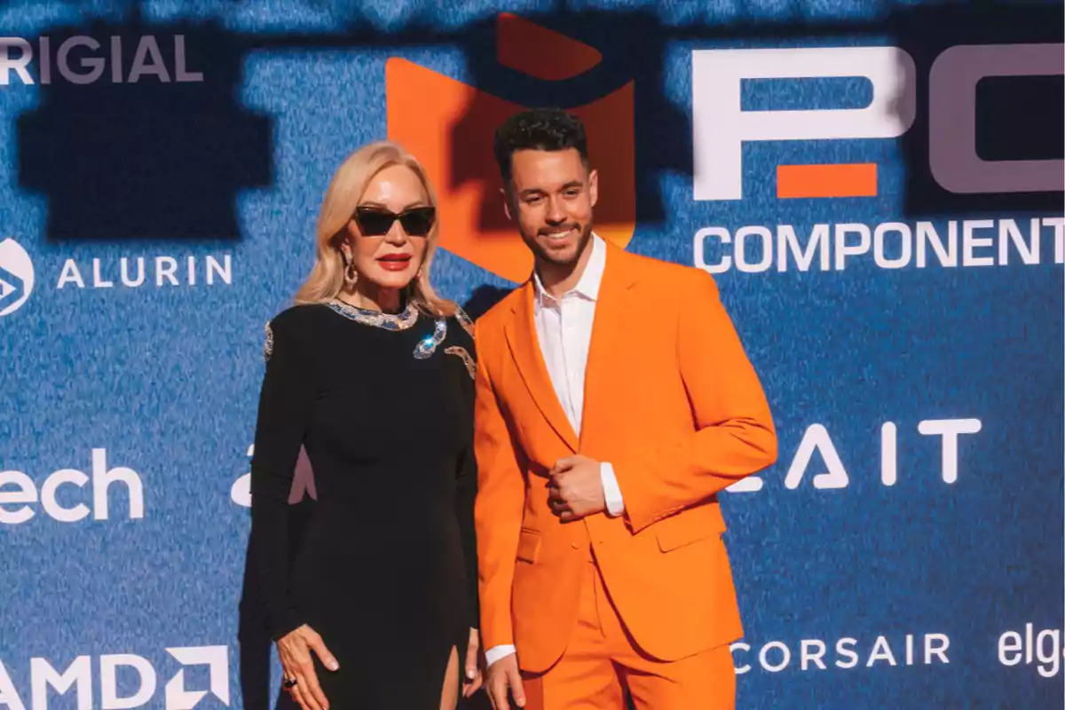 Dos personas posan frente a un fondo azul con logotipos y texto, una lleva un vestido negro y gafas de sol, y la otra un traje naranja.
