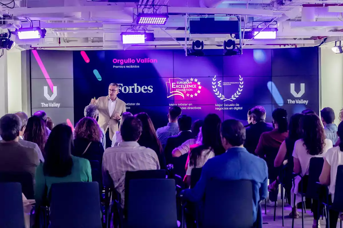 Un hombre de pie da una presentación frente a una audiencia en un auditorio moderno con pantallas que muestran logotipos de premios y reconocimientos de una escuela de negocios digitales llamada The Valley