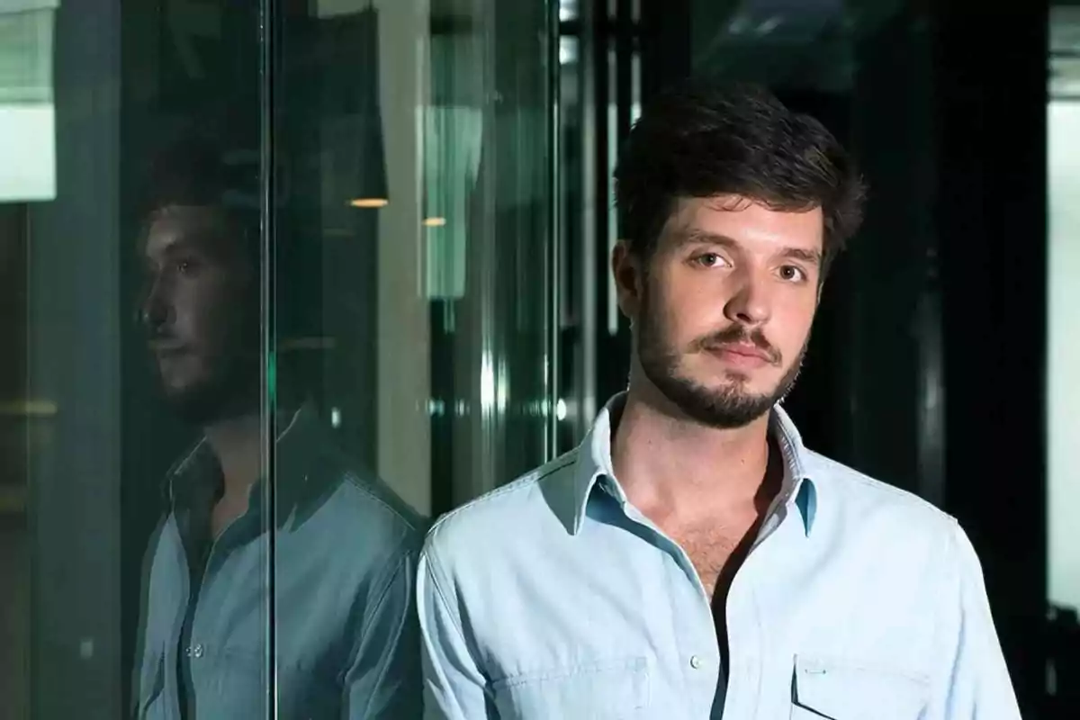 Un hombre con barba y camisa clara está de pie junto a una pared de vidrio en un entorno interior moderno.