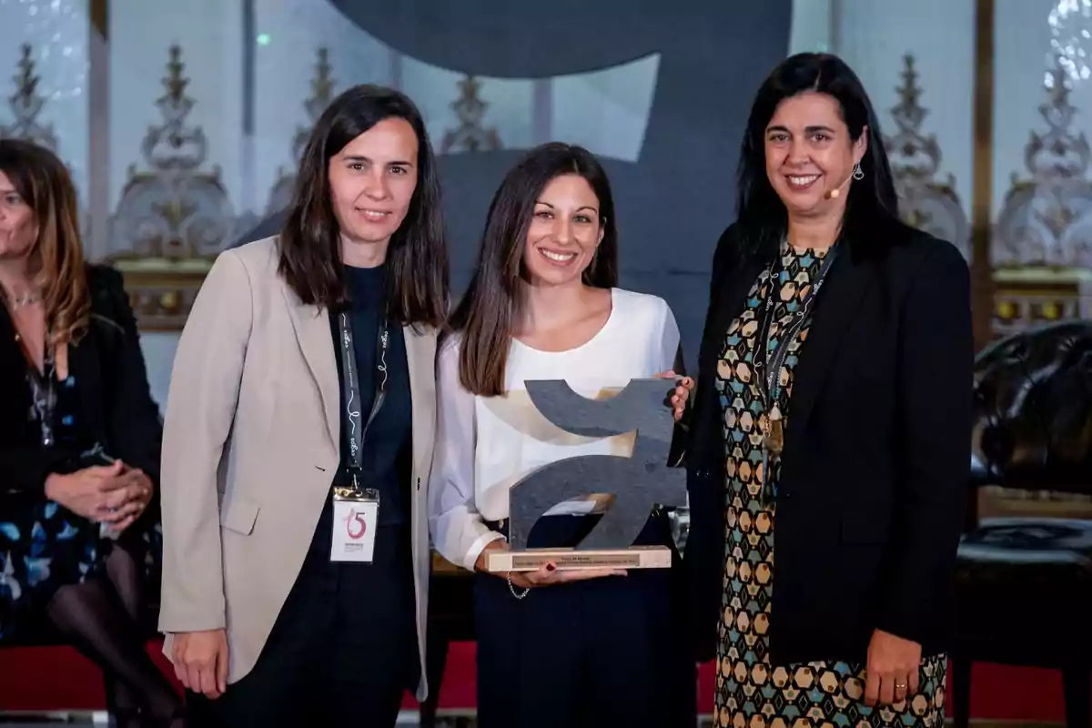 Tres mujeres posan juntas en un evento, una de ellas sostiene un trofeo.