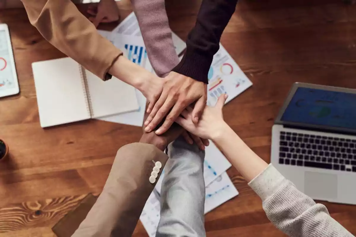 Un grupo de personas uniendo sus manos sobre una mesa de trabajo con documentos y una laptop.