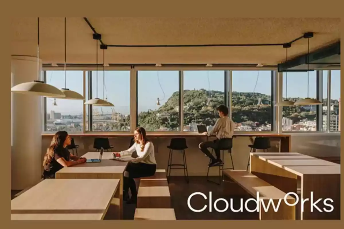 Personas trabajando en un espacio de oficina moderno con mesas de madera y grandes ventanales que ofrecen vistas a una ciudad y colinas al fondo.