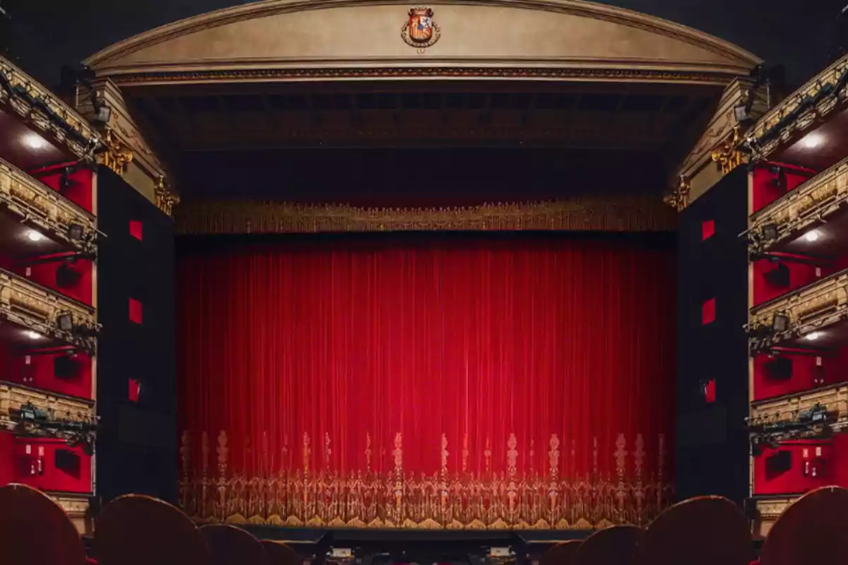 Un teatro con un gran telón rojo cerrado y balcones decorados a los lados.