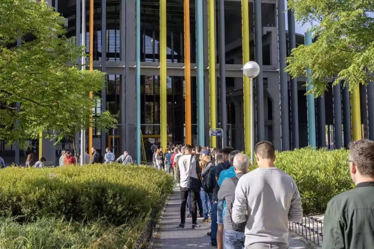 Personas haciendo fila frente a un edificio con columnas de colores y rodeado de vegetación.