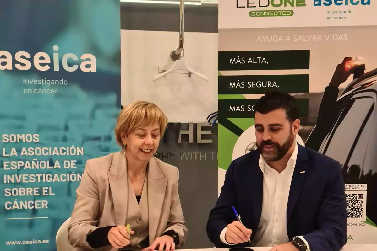 Dos personas firmando documentos en un evento de colaboración entre Aseica y LedOne, con pancartas de fondo que destacan la investigación en cáncer y la seguridad.