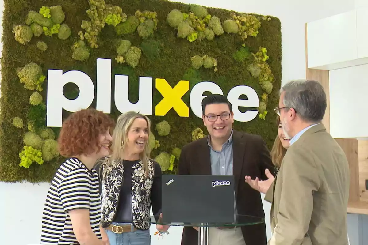 Un grupo de personas sonríe y conversa frente a un letrero de "pluxee" con un fondo de musgo verde.