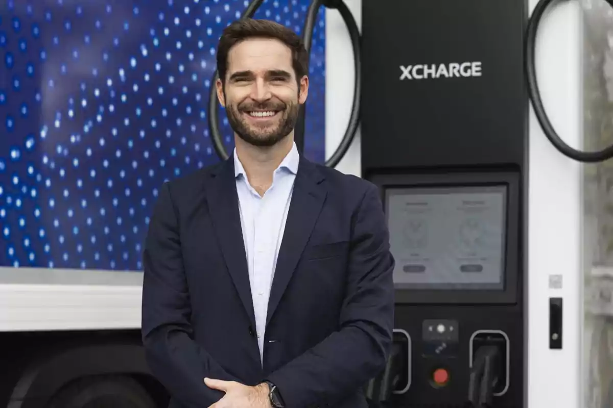 Hombre sonriendo frente a una estación de carga eléctrica con fondo azul.