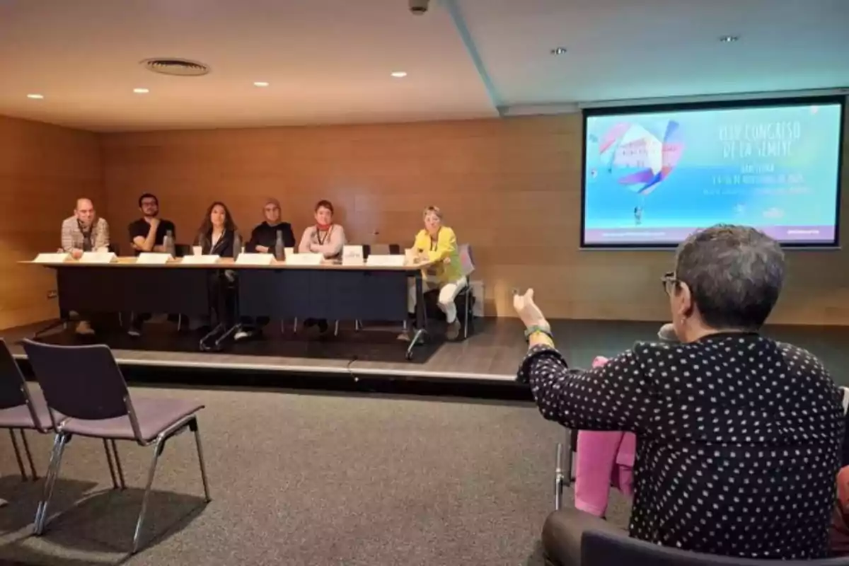 Un panel de seis personas está sentado en una mesa frente a una audiencia en una sala de conferencias, mientras una persona del público hace una pregunta.