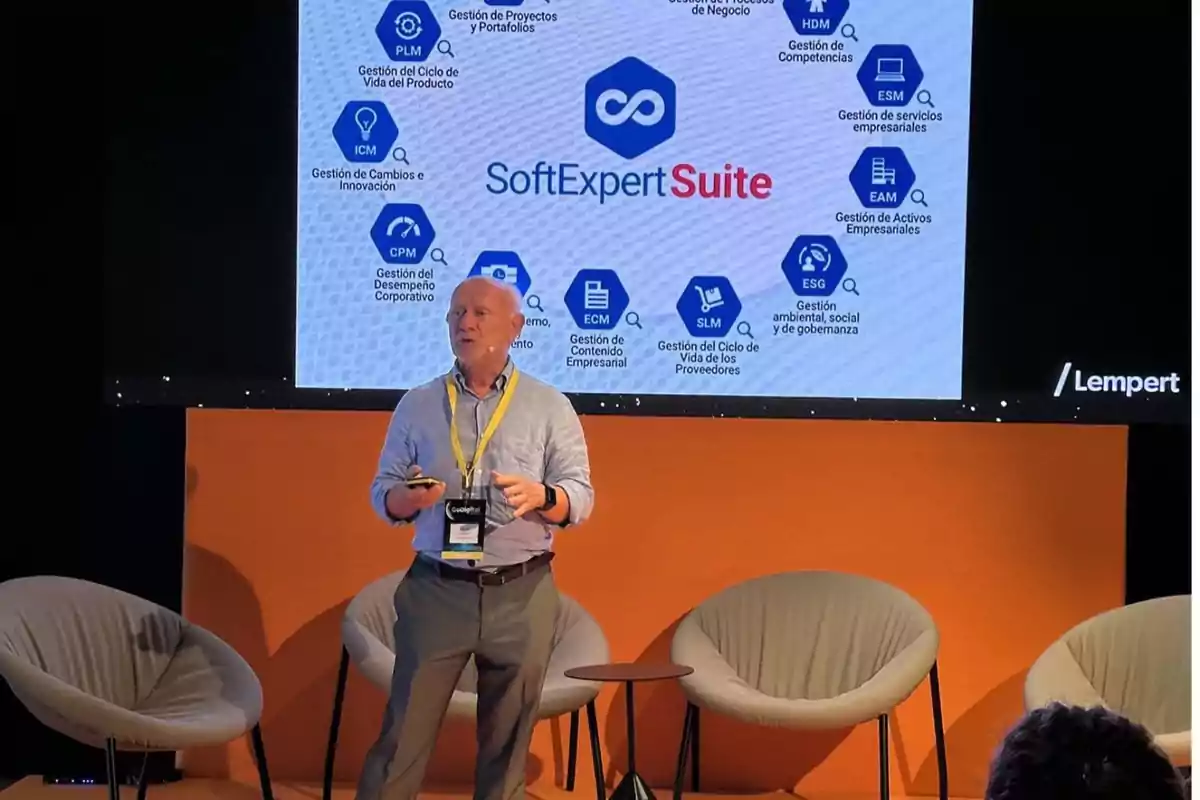 Un hombre presenta SoftExpert Suite en un escenario con sillas y una pantalla que muestra diferentes áreas de gestión empresarial.