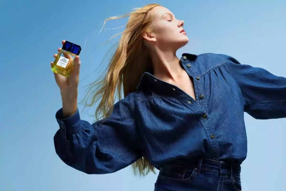 Mujer con camisa de mezclilla sosteniendo un frasco de perfume amarillo con tapa azul sobre un fondo azul claro.