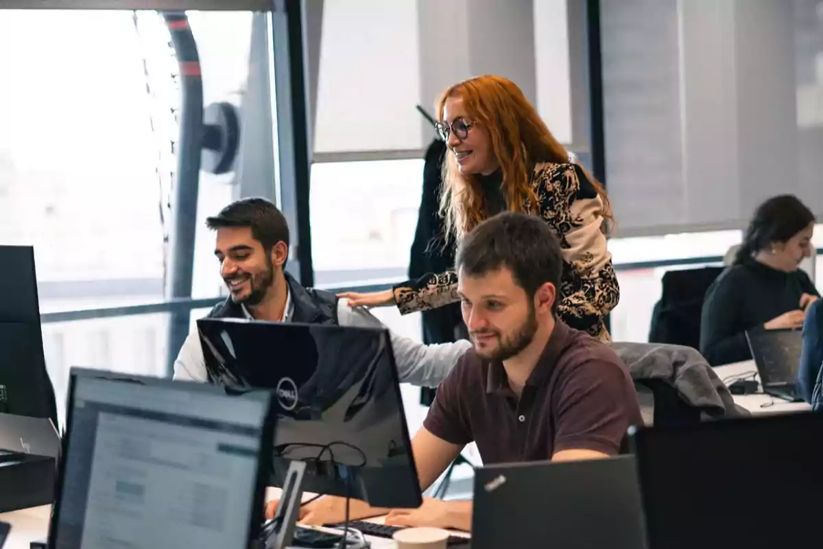 Personas trabajando en una oficina moderna con computadoras y ventanas grandes.