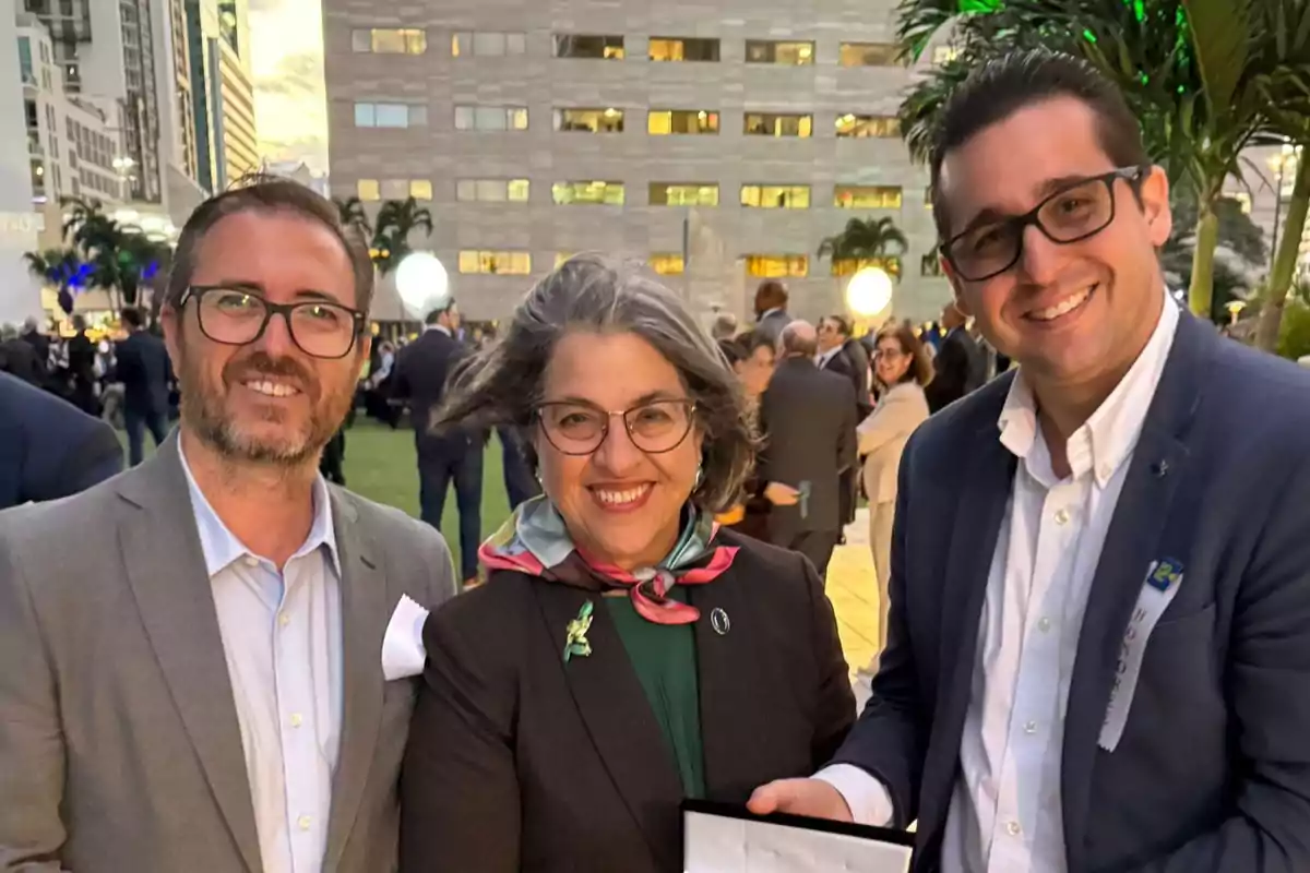 Tres personas sonríen a la cámara en un evento al aire libre con edificios y palmeras de fondo.