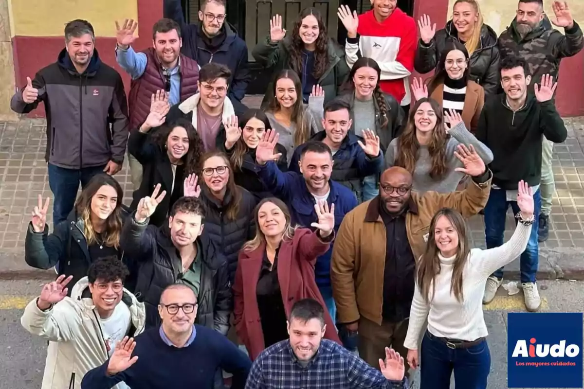 Un grupo de personas sonrientes levanta la mano en señal de saludo mientras posan para una foto en la calle.