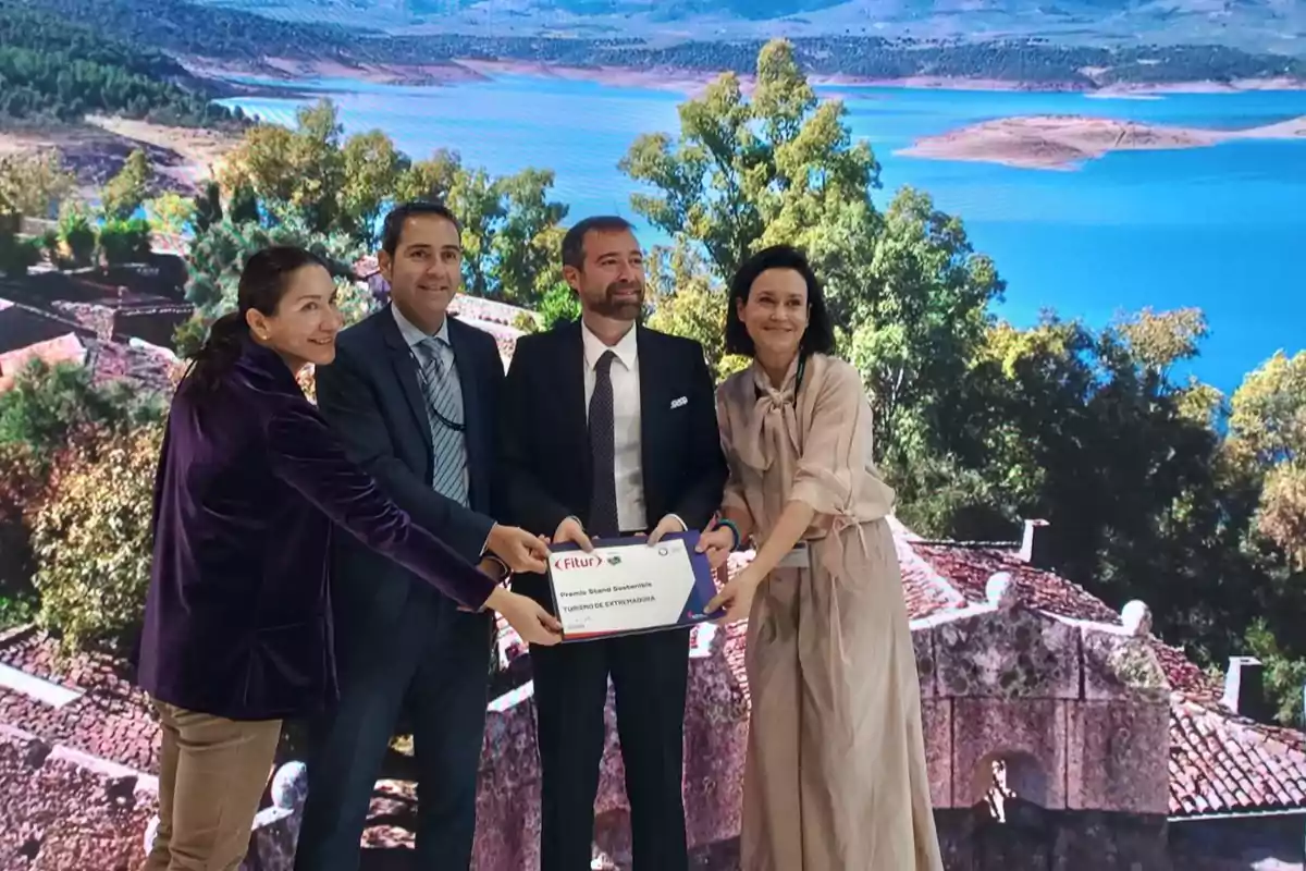 Cuatro personas posan juntas sosteniendo un cartel de Fitur frente a un fondo de paisaje con árboles y un lago.