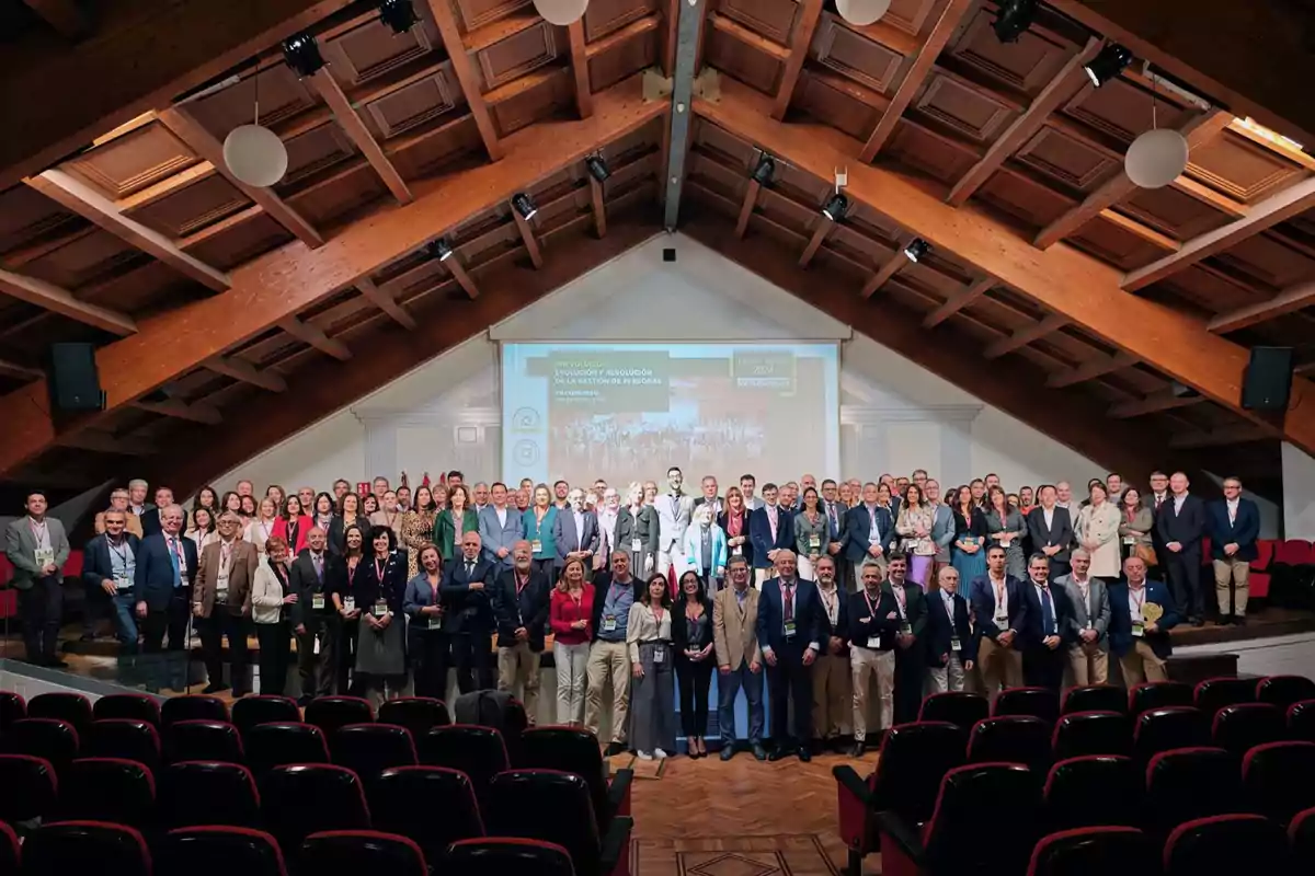 Un grupo numeroso de personas posando en un auditorio con un techo de madera y una pantalla de proyección al fondo.