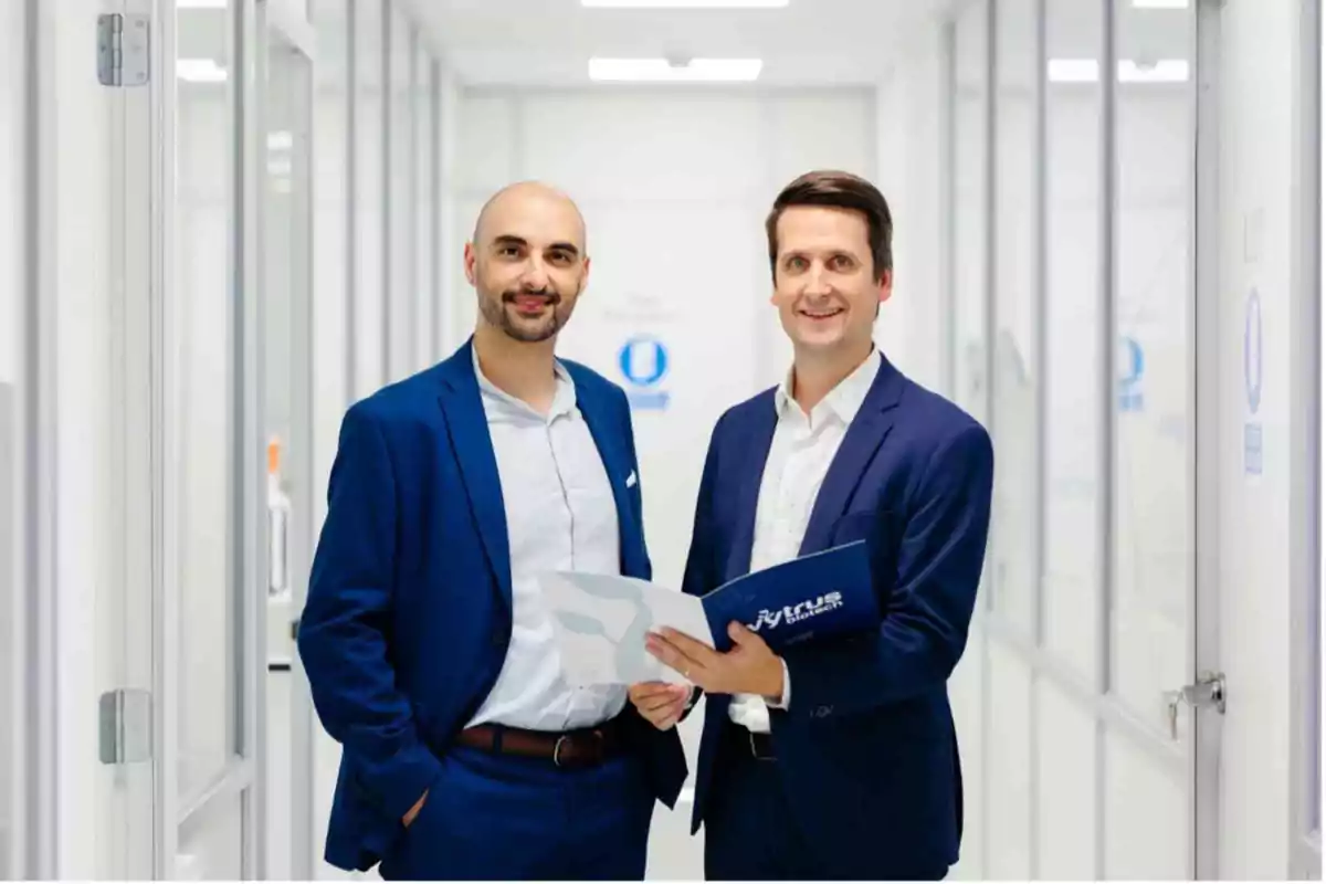 Dos hombres de traje azul posan en un pasillo de oficina mientras sostienen documentos.