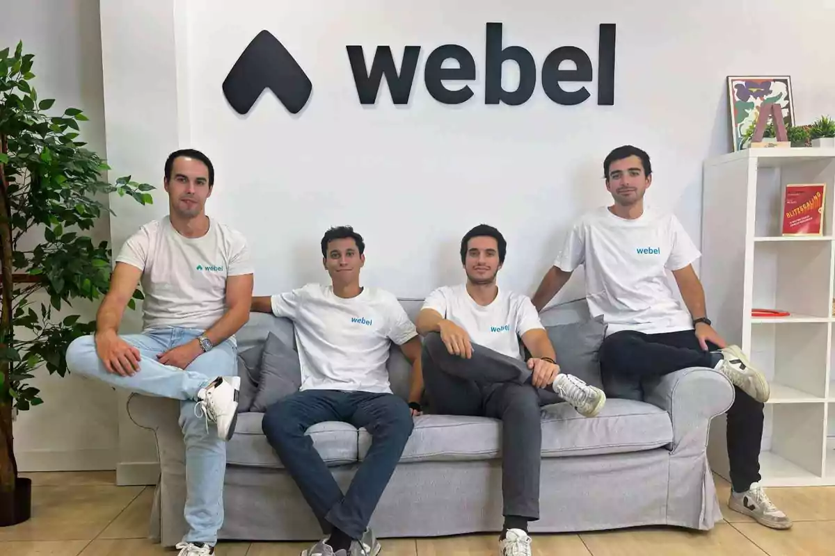 Cuatro hombres sentados en un sofá con camisetas blancas que tienen el logo de "webel" en una oficina moderna.