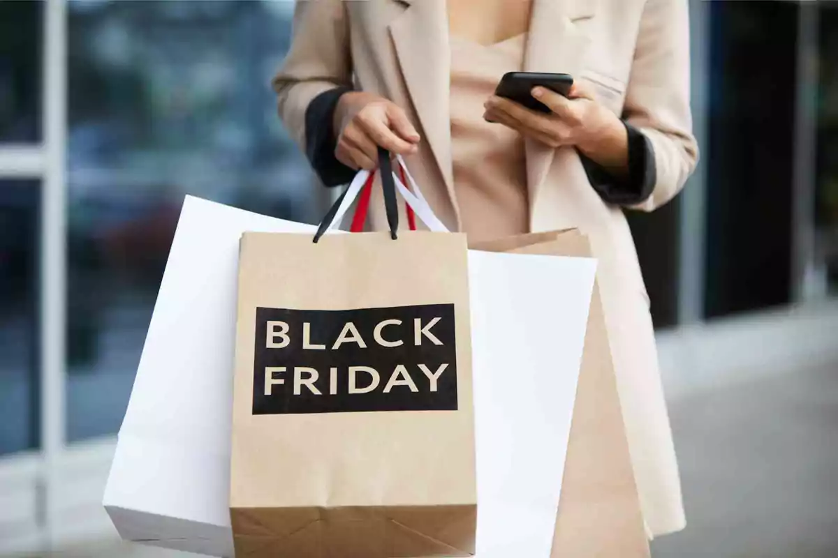 Persona sosteniendo bolsas de compras con la etiqueta Black Friday mientras revisa su teléfono móvil.