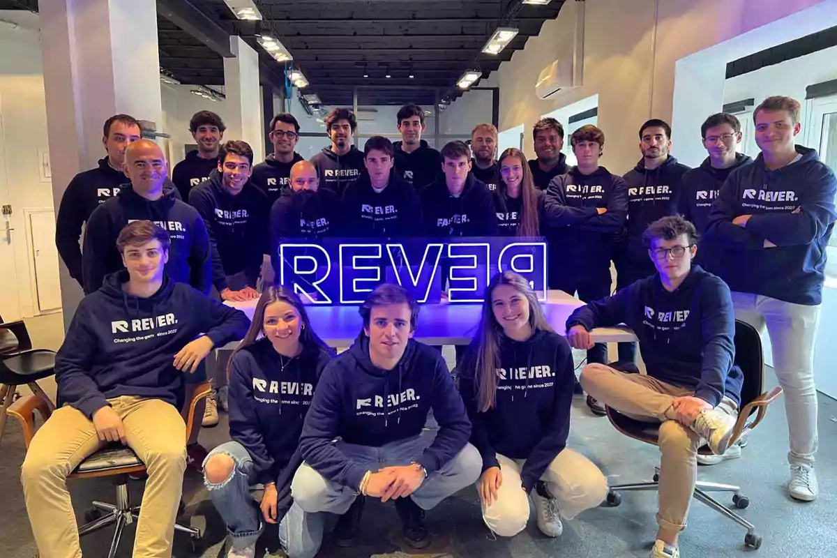 Un grupo de personas con sudaderas de "REVER" posando alrededor de un letrero iluminado en una oficina.