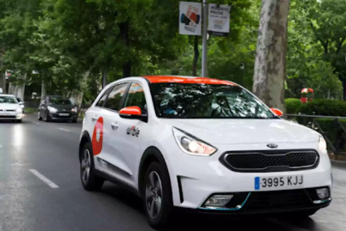 Un coche blanco de alquiler circula por una calle arbolada.