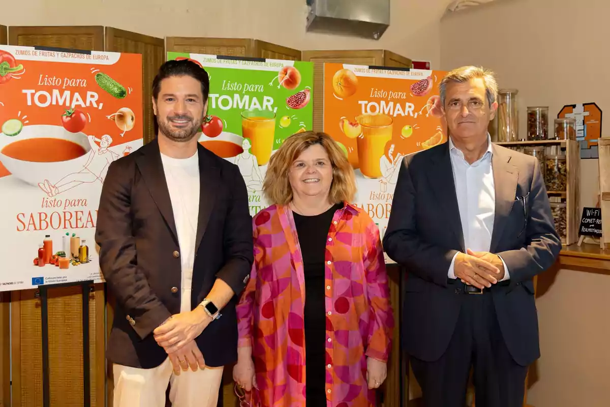 Tres personas posan sonrientes frente a carteles promocionales de zumos y gazpachos en un evento interior