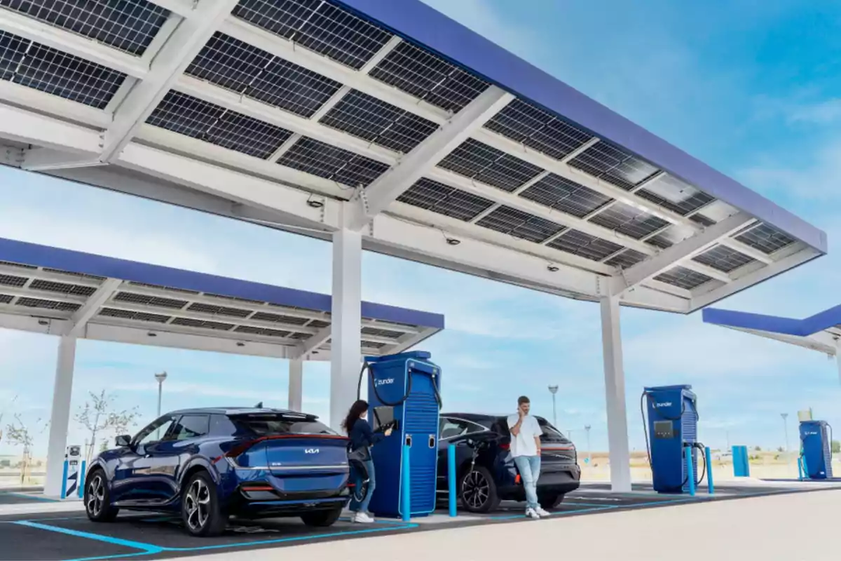 Estación de carga para vehículos eléctricos con paneles solares en el techo y dos personas utilizando los cargadores.